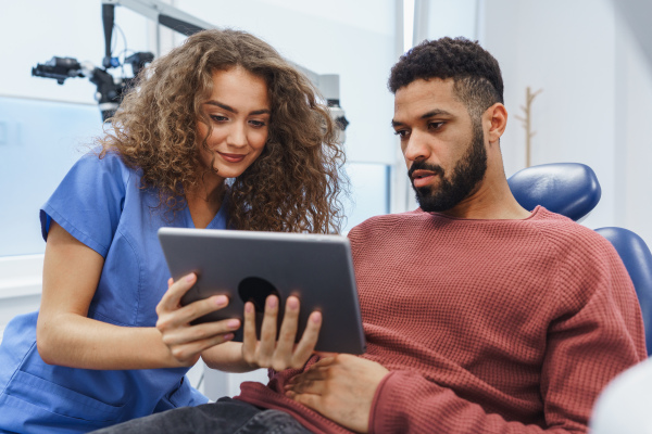 Young woman dentist doing preventive examination to multiracial man, showing him x-ray image in a digital tablet.