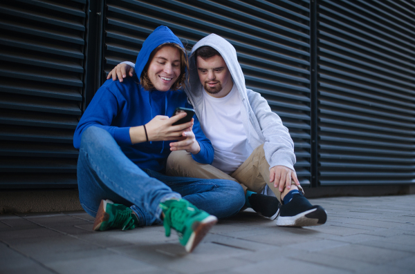 Man with down syndrome resting outdoor with his friend, scrolling mobile phone. Concept of friendship and integration people with disability into society.