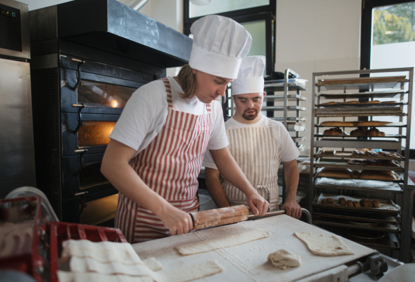 Man with down syndrom helping prepair bread in a bakery with his colleague. Concept of integration people with disability into society.