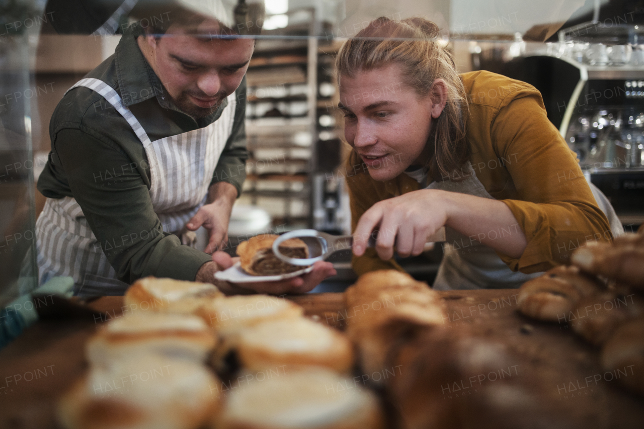 Man with down syndrom helping his colleague in a cafe, carring fresh pastries. Concept of integration people with a disabilities into society.