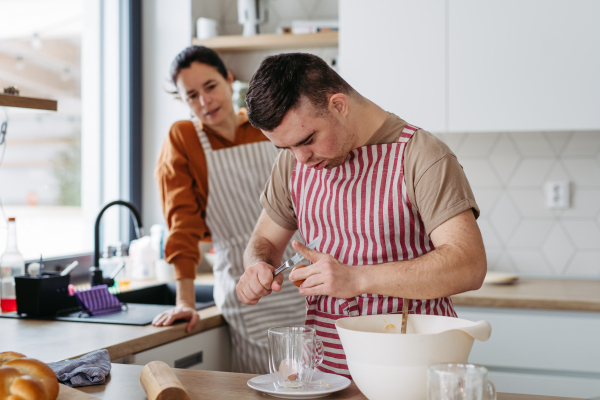 Young man with Down syndrome preparing breakfast with his mother at home, cracking eggs. Morning routine for man with Down syndrome genetic disorder.