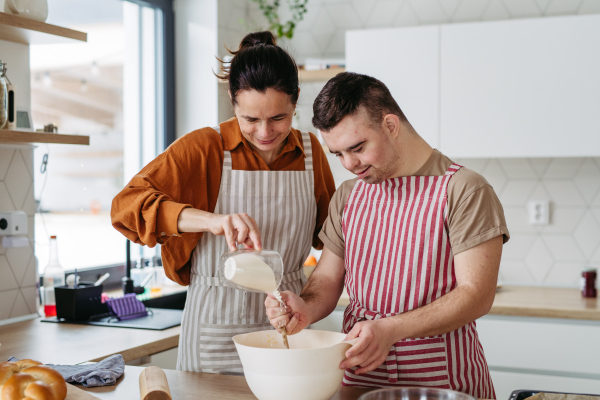 Young man with Down syndrome preparing breakfast with his mother at home, cracking eggs. Morning routine for man with Down syndrome genetic disorder.