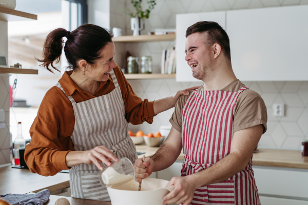 Young man with Down syndrome preparing breakfast with his mother at home, cracking eggs. Morning routine for man with Down syndrome genetic disorder.