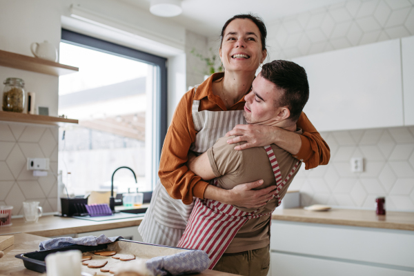 Young man with Down syndrome baking cookies, sweets with his mother at home, hugging her. Daily routine for man with Down syndrome. Concept of mothers day.