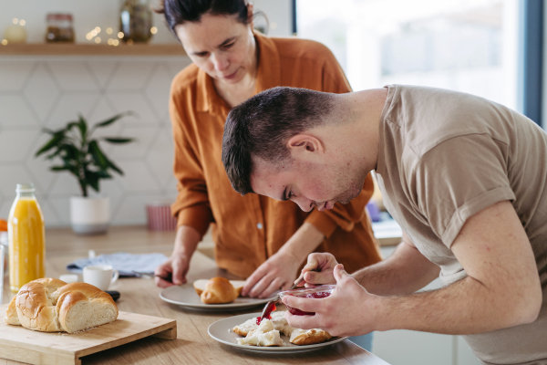 Young man with Down syndrome preparing breakfast with his mother at home. Morning routine for man with Down syndrome genetic disorder.