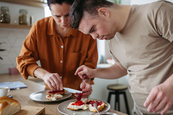 Young man with Down syndrome preparing breakfast with his mother at home. Morning routine for man with Down syndrome genetic disorder.