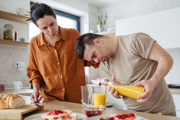 Young man with Down syndrome pouring juice, preparing breakfast with his mother at home. Morning routine for man with Down syndrome genetic disorder.