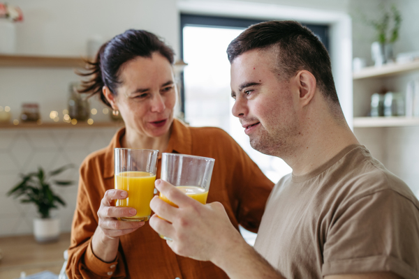 Portrait of young man with Down syndrome with his mother at home, toasting with juice. Morning routine for man with Down syndrome genetic disorder.