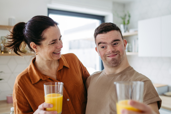 Portrait of young man with Down syndrome with his mother at home, toasting with juice. Morning routine for man with Down syndrome genetic disorder.