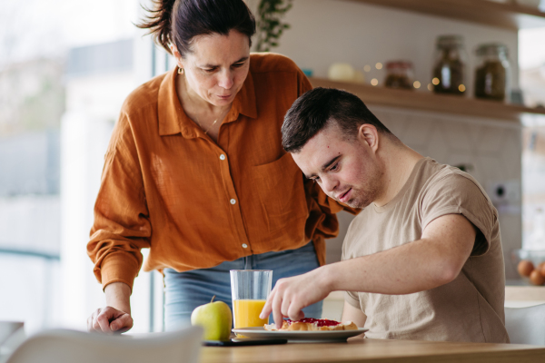 Young man with Down syndrome preparing breakfast with his mother at home. Morning routine for man with Down syndrome genetic disorder.