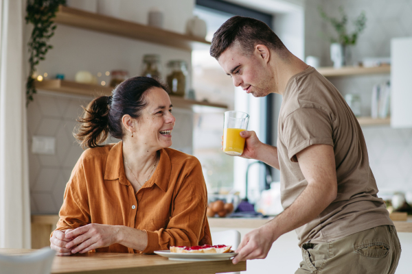 Portrait of young man with Down syndrome serving breakfast to his mother as a Mother's Day gift. Concept of Mother's Day and maternal love to disabled children.