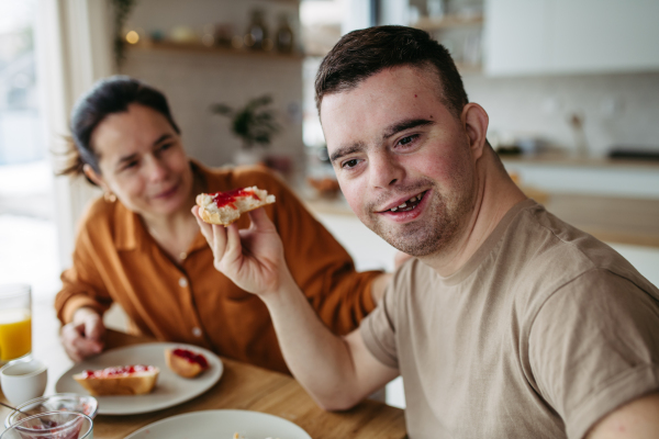 Young man with Down syndrome having breakfast with his mother at home. Morning routine for man with Down syndrome genetic disorder.