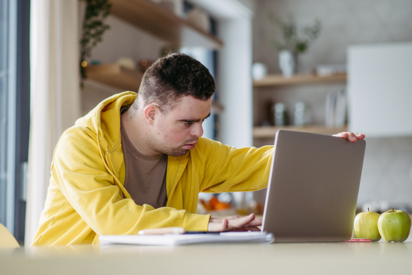 Young man with down syndrome learning online at home, using laptop, working on notebook. Telehealth consultation with doctor.