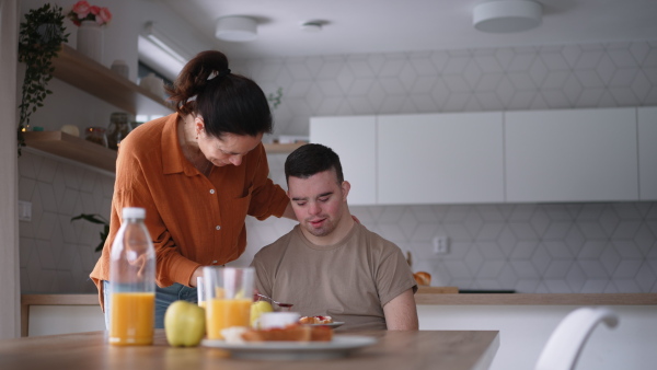 Young man with Down syndrome having breakfast with his mother at home. Morning routine for man with Down syndrome genetic disorder.