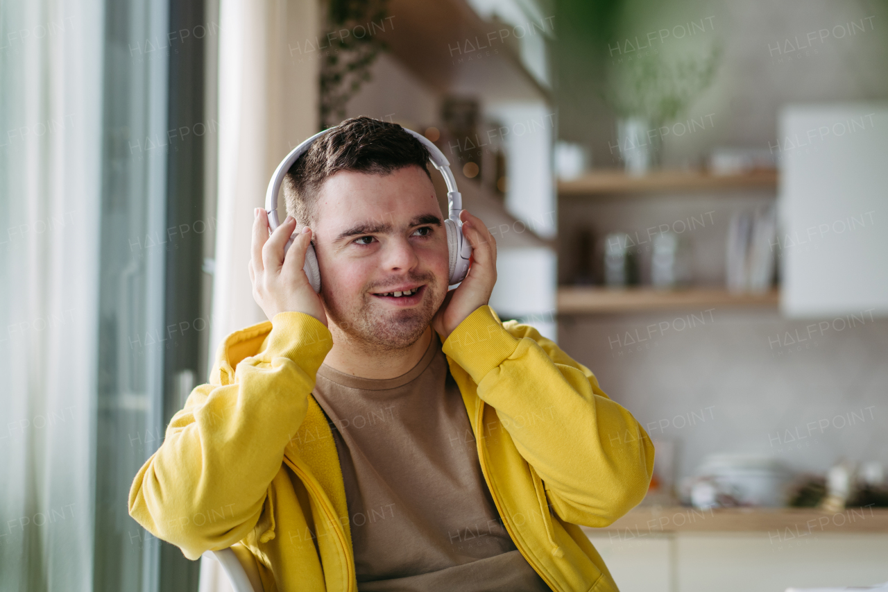 Young man with down syndrome listening his music via headphones. Sitting in the kitchen, wireless headphones on head.