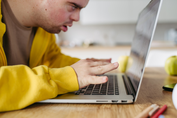 Young man with down syndrome learning online at home, using laptop, working on notebook. Telehealth consultation with doctor.