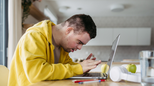 Young man with down syndrome learning online at home, using laptop, working on notebook. Telehealth consultation with doctor.