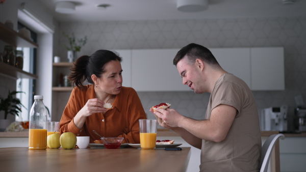 Portrait of young man with Down syndrome with his mother at home, toasting with juice. Morning routine for man with Down syndrome genetic disorder.