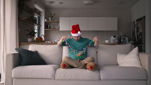 Young man with Down syndrome and santa hat sitting on sofa playing with christmas lights.