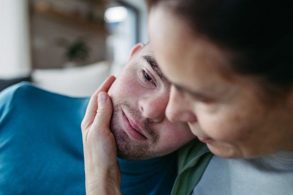 Portrait of young man with Down syndrome with his mother at home, hugging an stroking cheek. Concept of love and parenting disabled child.