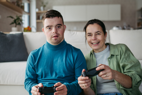 Portrait of young man with Down syndrome with his mother playing video game, holding game controllers. Concept of love and parenting disabled child.