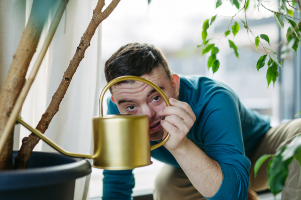 Young man with Down syndrome taking care of indoor plants, watering them, looking at the camera through handle of a metal watering can.