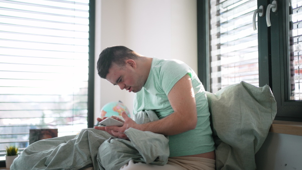 Young man with down syndrome lying in his bed, looking at smartphone in the morning. Morning routine for man with disability.