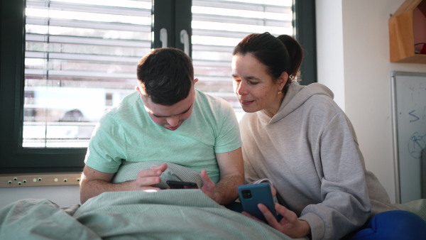 Young man with down syndrome sitting in his bed with mom, both looking at smartphone in the morning. Morning routine for man with disability.