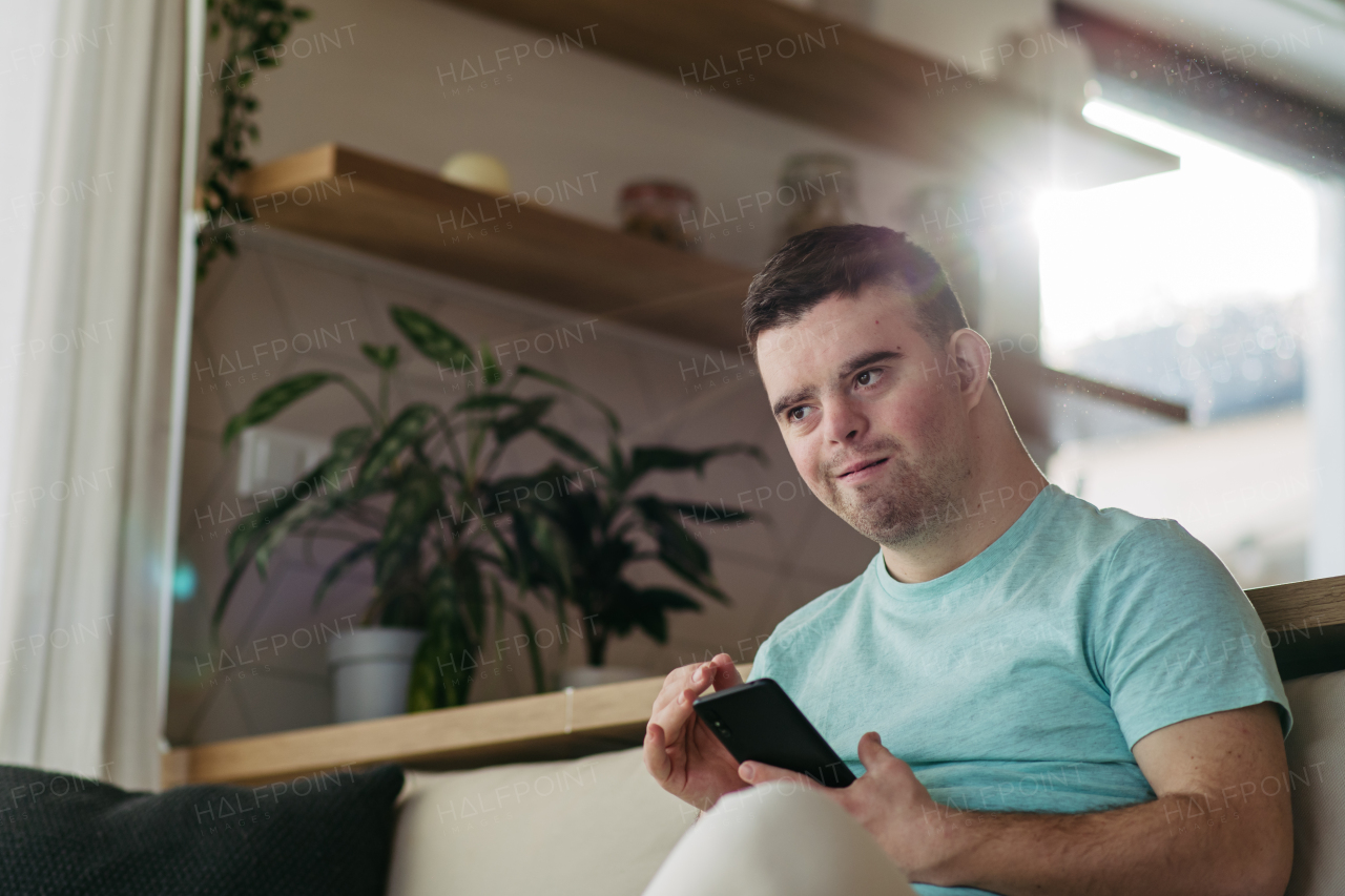 Portrait of young man with Down syndrome sitting in kitchen with smartphone in hand.