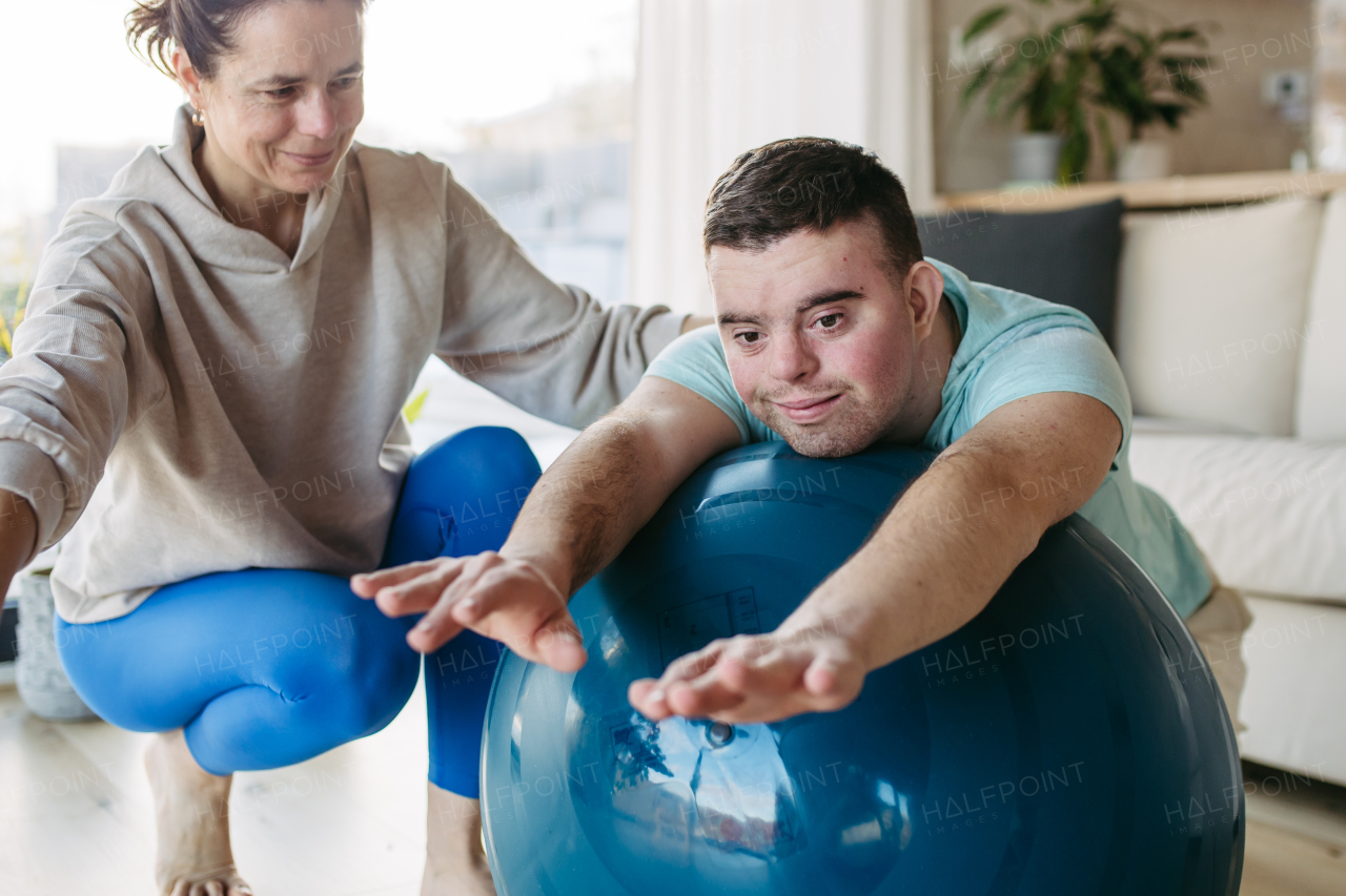 Young man with Down syndrome exercising at home with his mother on a fitness ball. Workout routine for disabled man.