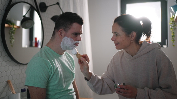 Mother applying shaving foam on son's face. Young man with down syndrome learning how to shave. Concept of mother's day and motherly love.