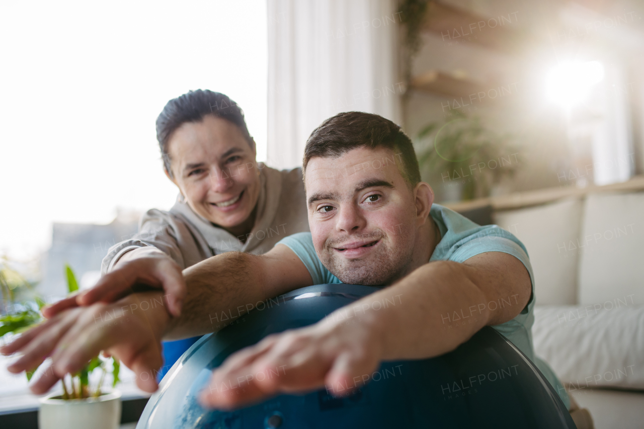 Young man with Down syndrome exercising at home with his mother on a fitness ball. Workout routine for disabled man.