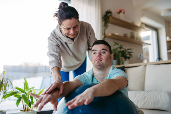 Young man with Down syndrome exercising at home with his mother on a fitness ball. Workout routine for disabled man.