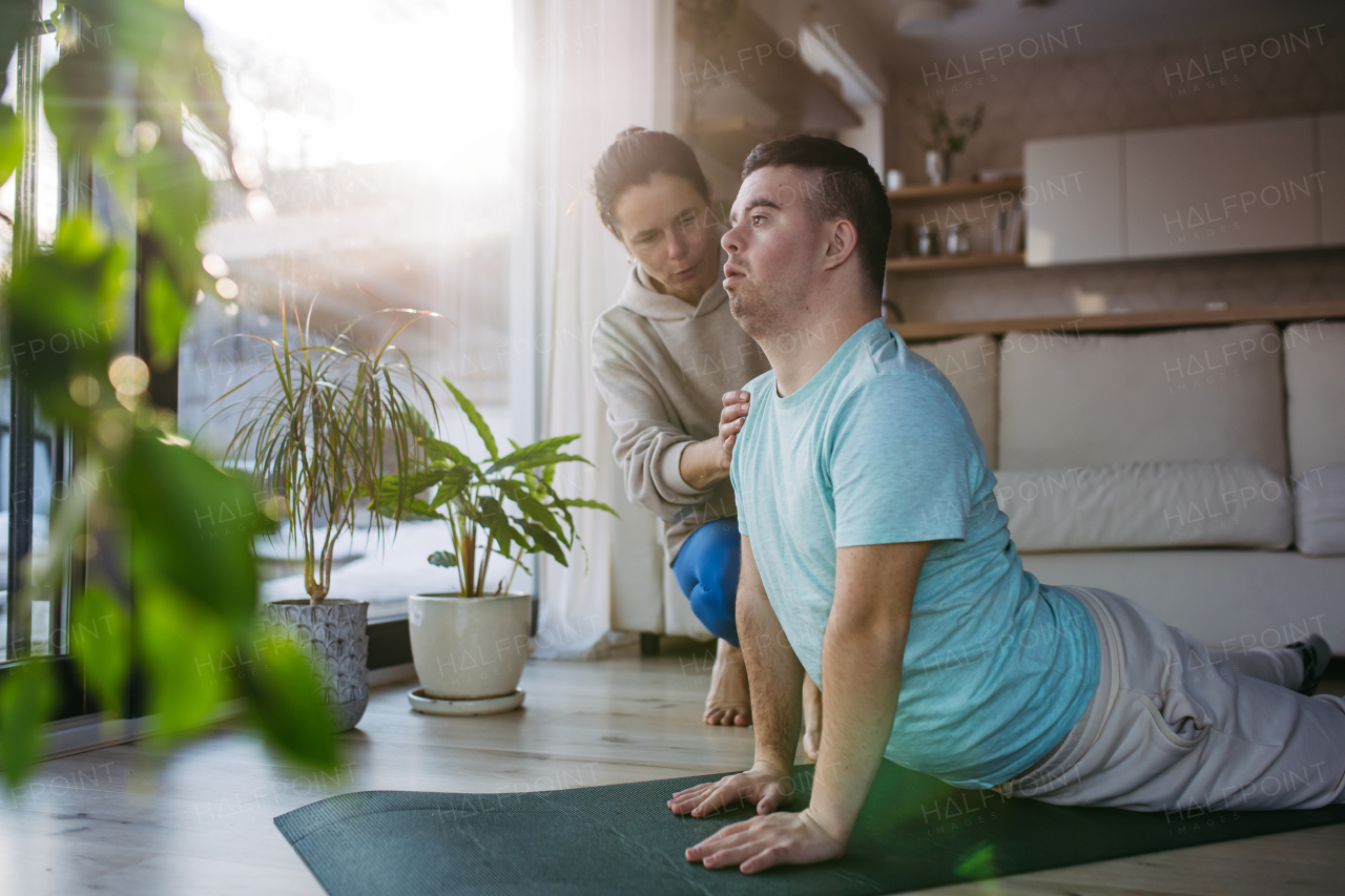 Young man with Down syndrome exercising at home with his mother on a fitness ball. Workout routine for disabled man.