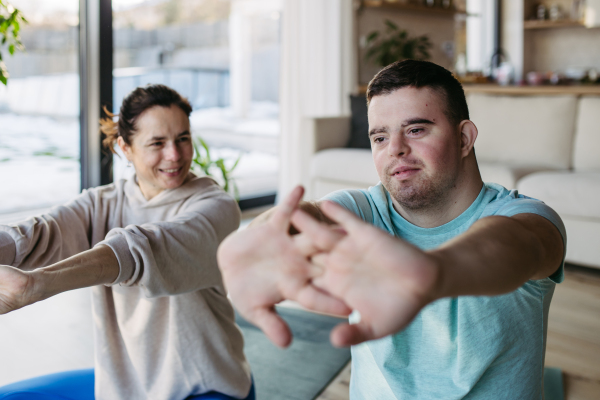 Young man with Down syndrome exercising at home with his mother on a fitness ball. Workout routine for disabled man.