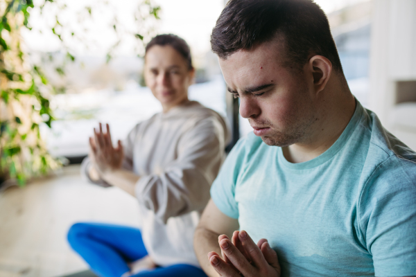 Young man with Down syndrome exercising at home with his mother on gym matroutine for disabled man.