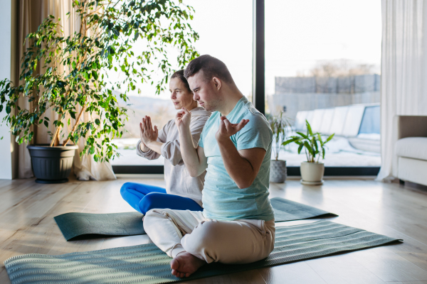 Young man with Down syndrome doing yoga at home with his mother on a fitness ball. Workout routine for disabled man.