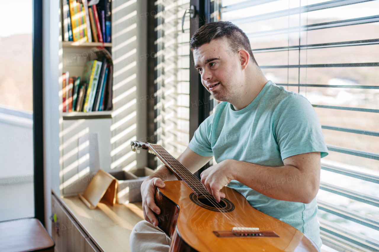 Young man with down syndrome playing acoustic guitar, sitting by window, holding and strumming a guitar, making beautiful music.