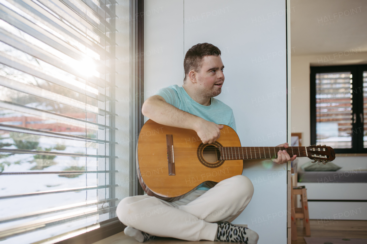 Young man with down syndrome playing acoustic guitar, sitting by window, holding and strumming a guitar, making beautiful music.