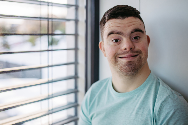 Portrait of young man with Down syndrome sitting by window, looking at camera, smiling.