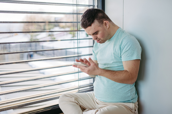 Portrait of young man with Down syndrome sitting by window with smartphone in hand and scrolling.