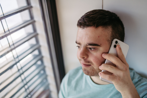 Portrait of young man with Down syndrome sitting by window, making phonecall with smartphone.