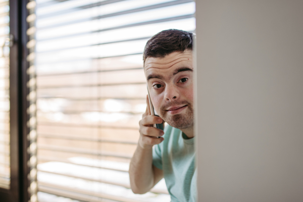 Portrait of young man with Down syndrome sitting by window, making phone call with smartphone.