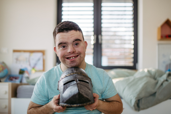 Portrait of happy smiling man with down syndrome at home, holding knight's helmet.