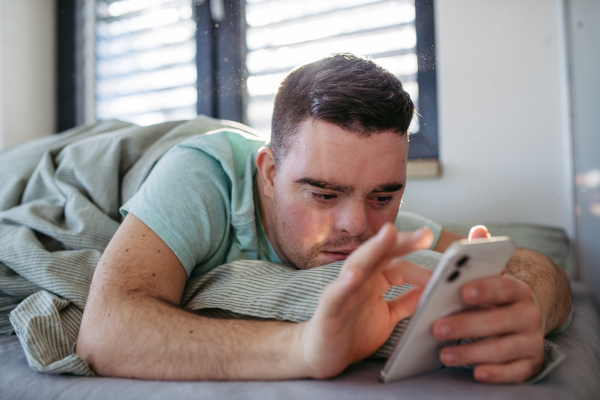 Young man with down syndrome lying in his bed, looking at smartphone in the morning. Morning routine for man with disability.