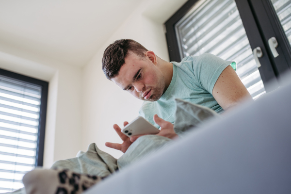 Young man with down syndrome sitting in his bed, looking at smartphone in the morning. Morning routine for man with disability.