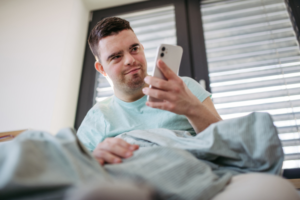 Young man with down syndrome lying in his bed, looking at smartphone in the morning. Morning routine for man with disability.