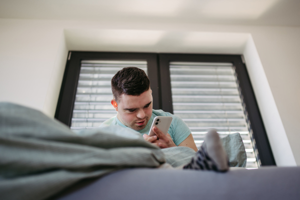 Young man with down syndrome lying in his bed, looking at smartphone in the morning. Morning routine for man with disability.