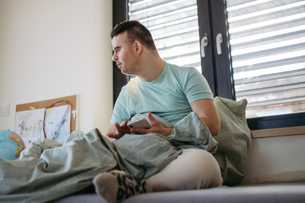 Young man with down syndrome sitting in his bed, looking at smartphone in the morning. Morning routine for man with disability.