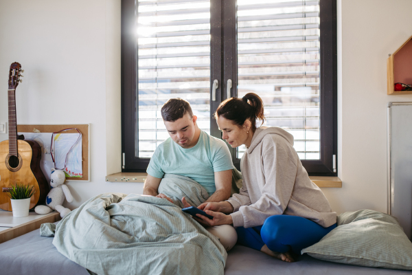 Young man with down syndrome sitting in his bed with mom, both looking at smartphone in the morning. Morning routine for man with disability.